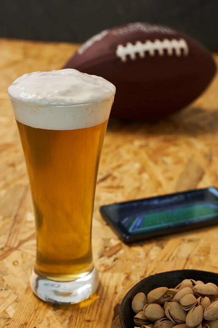 Hoher Blickwinkel auf ein Glas frisches, kaltes Bier auf einem Holztisch neben einem Teller mit Pistazien und einem Ball, bevor ein Spiel auf dem Handy angeschaut wird