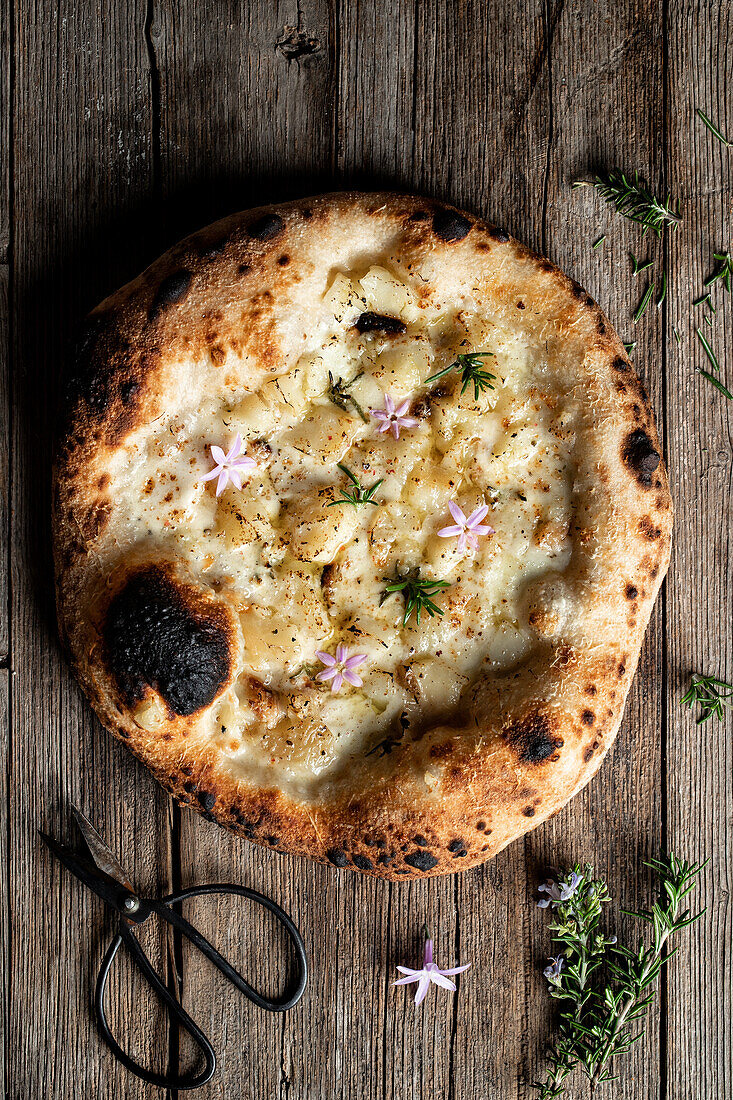 Overhead einer appetitlichen Pizza mit Käse, die mit essbaren kleinen Blumen und Rosmarin dekoriert ist und auf einem Holztisch liegt