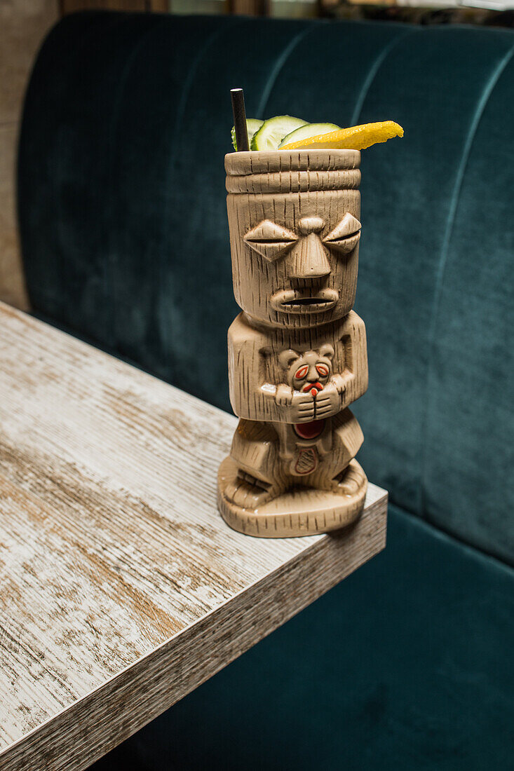 Traditional sculptural tiki cup of alcohol drink with straw placed on wooden table