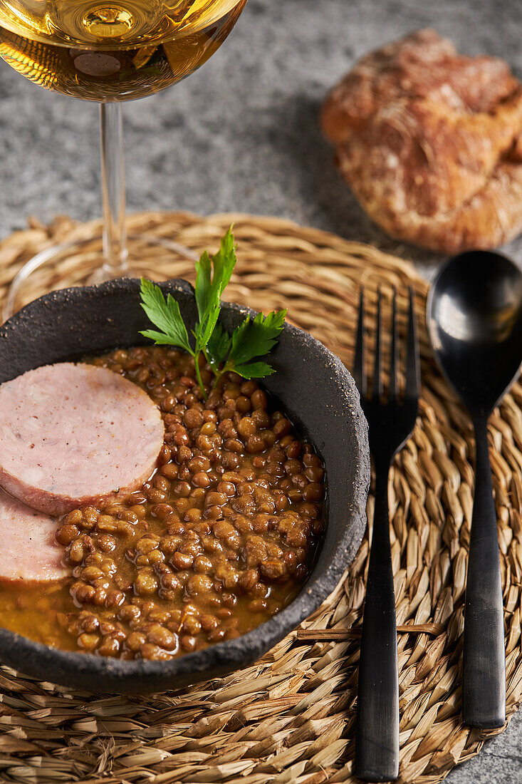 Top view rustic bowl with tasty lentil soup with parsley and slices of sausage placed on marble table near wine and cutlery during lunch