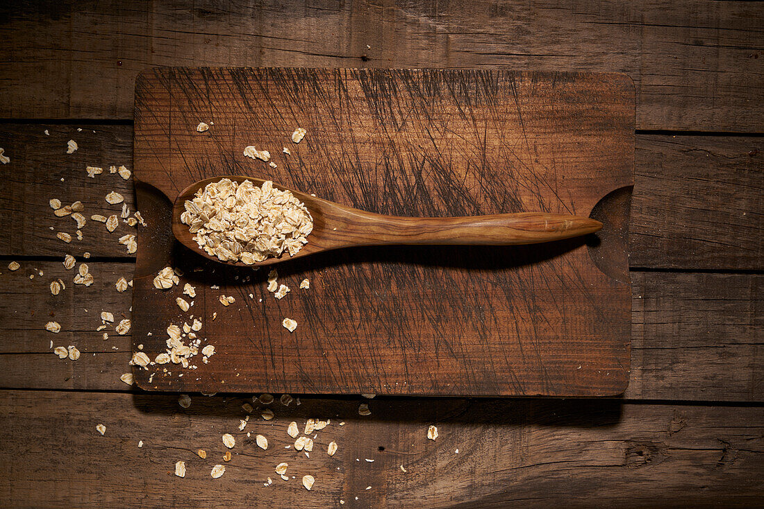Top view composition with wooden spoon filled with healthy oatmeal flakes placed on cutting board on rustic plank table