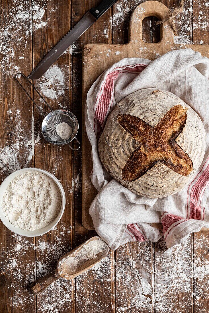 Draufsicht auf einen unordentlichen rustikalen Holztisch aus Weizenmehl und ein Brett mit frisch gebackenem Sauerteigbrotlaib