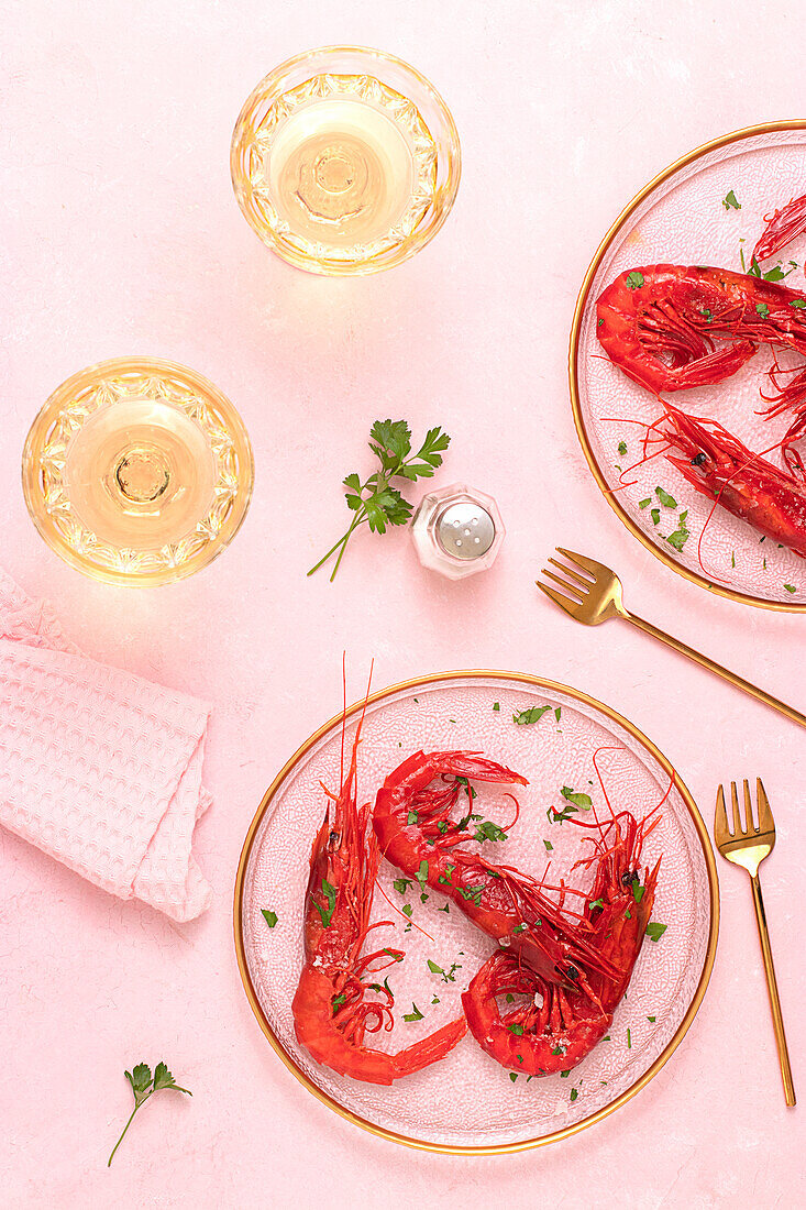 Blick von oben auf einen Glasteller mit leckeren gebratenen Tiger Shrimps neben einem Glas Wein auf einem Tisch mit rosa Hintergrund