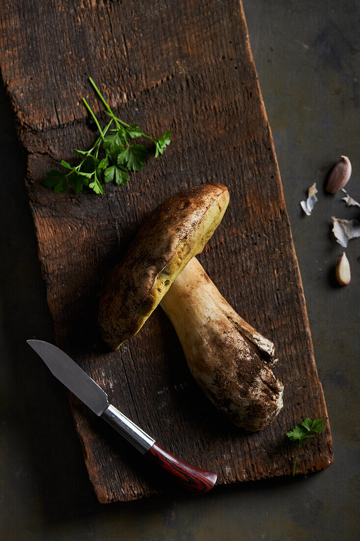 Draufsicht auf roh geschnittene Steinpilze (Boletus edulis) auf hölzernem Schneidebrett mit Knoblauch und Petersilie in heller Küche während des Kochvorgangs