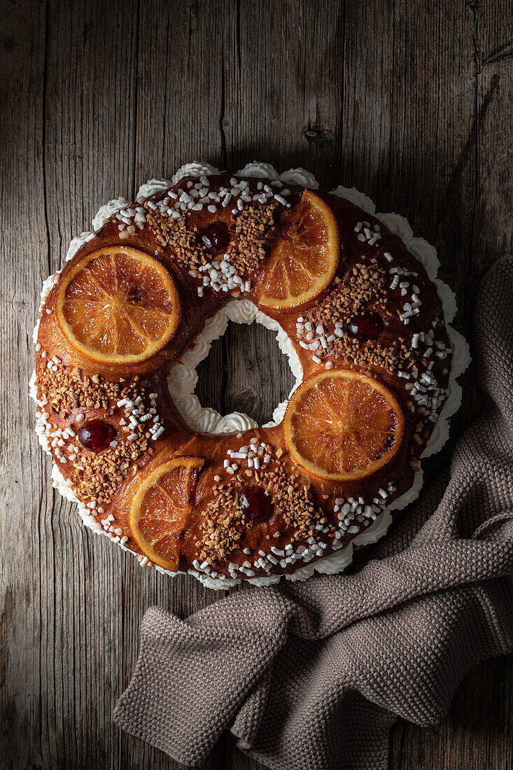 Overhead of king's cake with cream and sprinkles decorated with orange slices and cherries and placed on wooden surface near towel