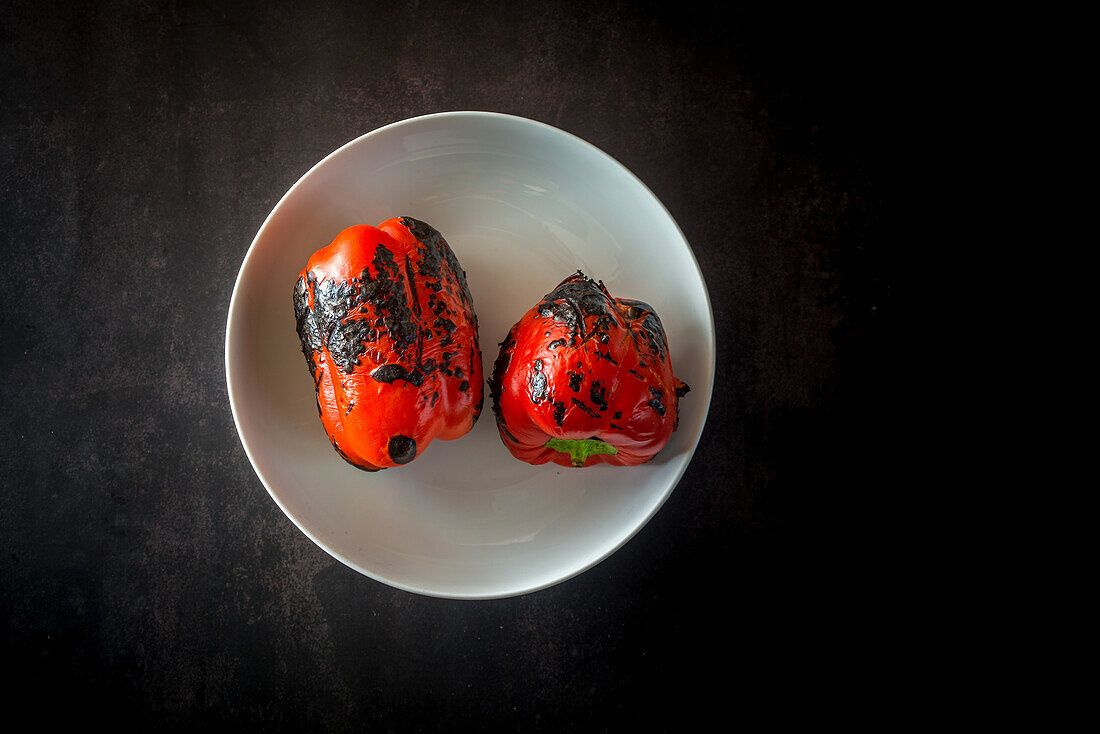 Draufsicht auf köstliche ganze gegrillte rote Paprika in einer runden Keramikschale auf dunklem Hintergrund