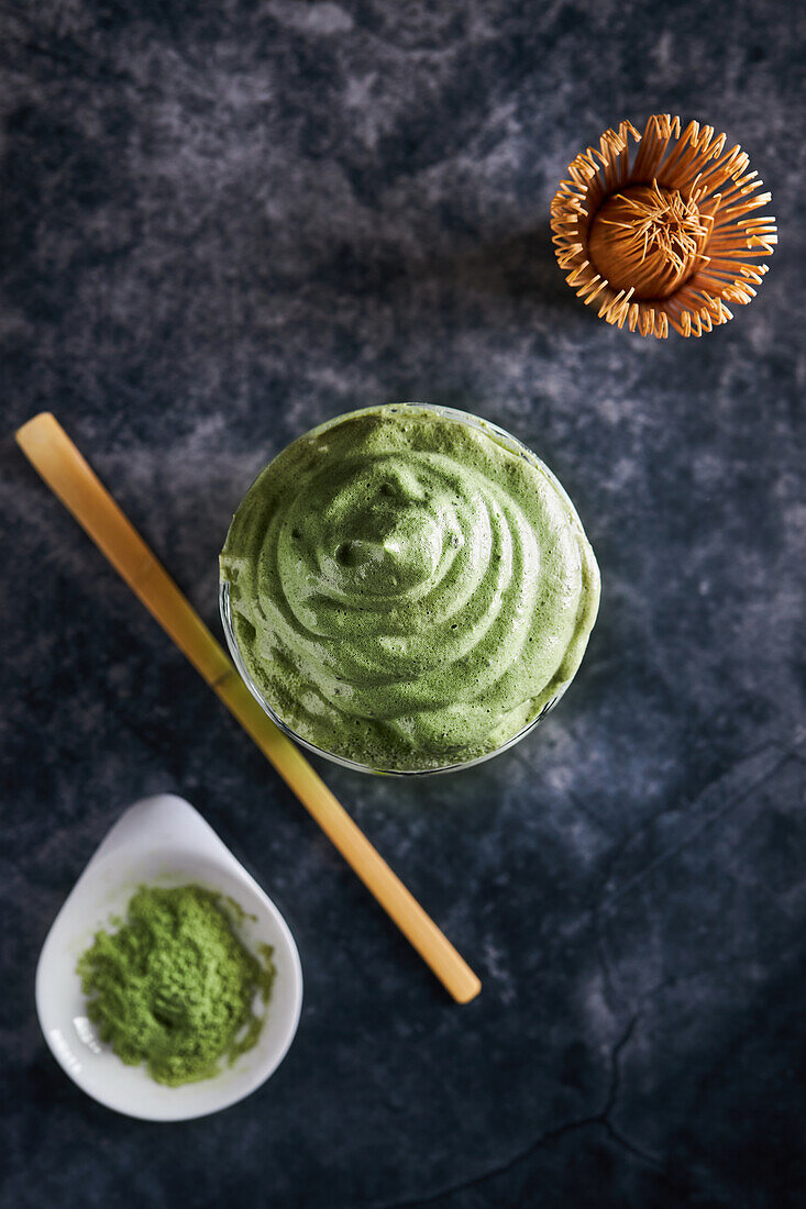 From above of glass of fresh dalgona coffee with matcha and Japanese tea ceremony accessories consisting of bamboo whisk called chasen and traditional scoop named chashaku placed on gray surface