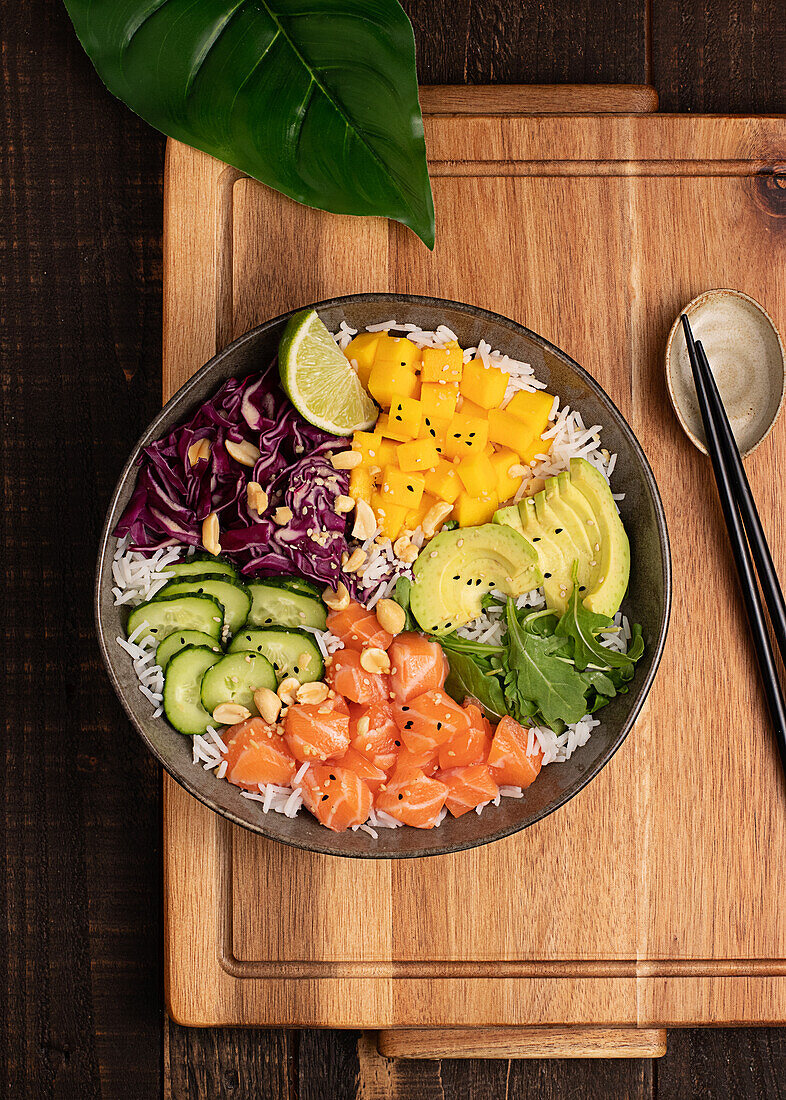 Top view of appetizing colorful poke bowl with fresh salmon and rice served with ripe avocado slices, cabbage and mango placed on wooden tray