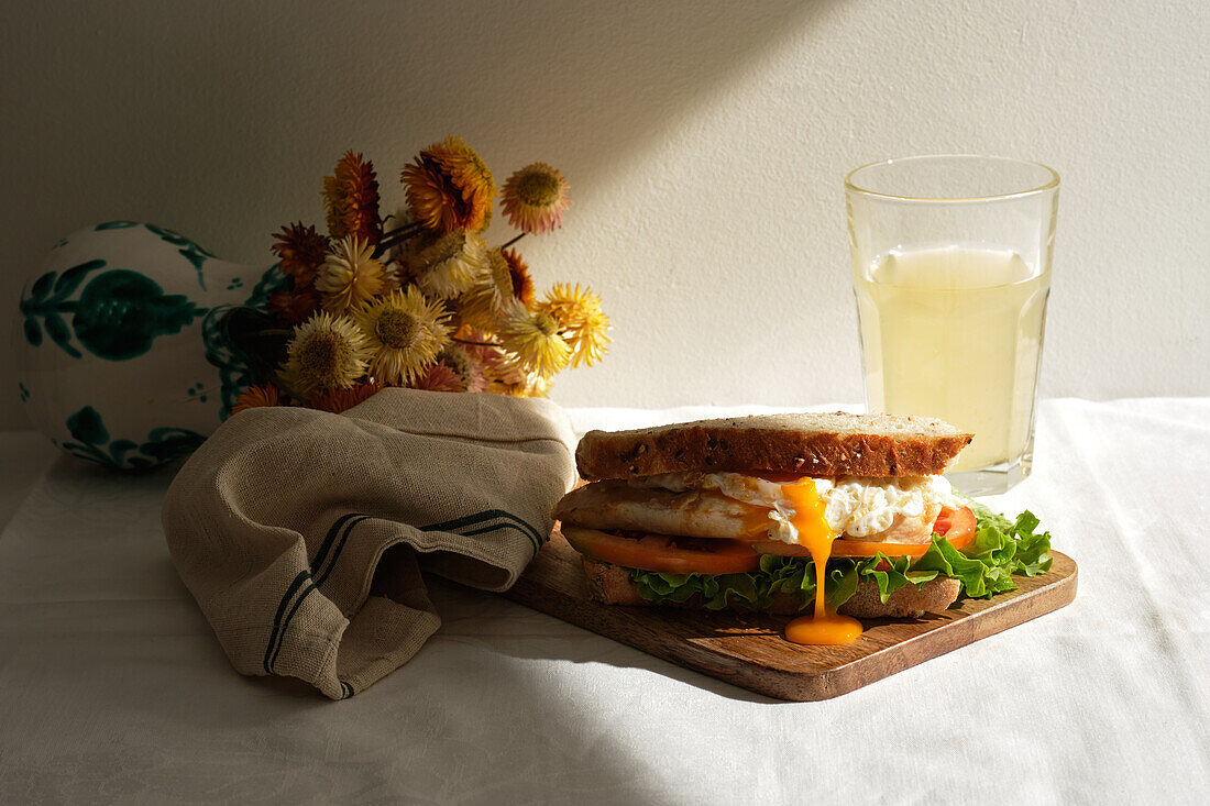 Appetitliches hausgemachtes Sandwich mit pochierten Eiern und frischen Tomaten und Salat, serviert mit einem Glas Saft zum Frühstück auf einem Tisch mit Blumen