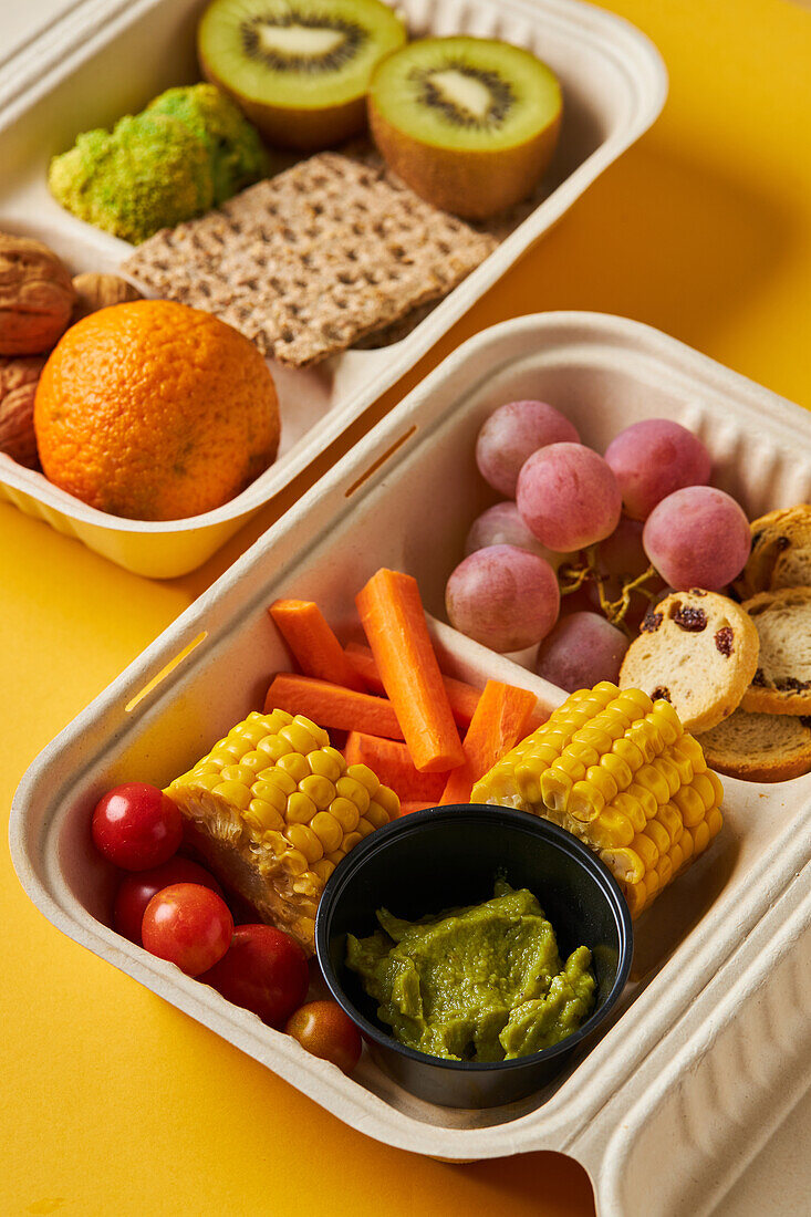 From above of lunch boxes with healthy food including crackers carrot sticks grapes cherry tomatoes with kiwi broccoli walnut and tangerine on yellow background