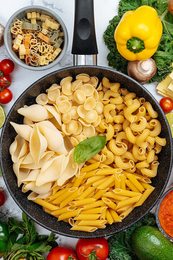 From above of frying pan with various uncooked pasta surrounded with assorted raw vegetables on marble table in light kitchen