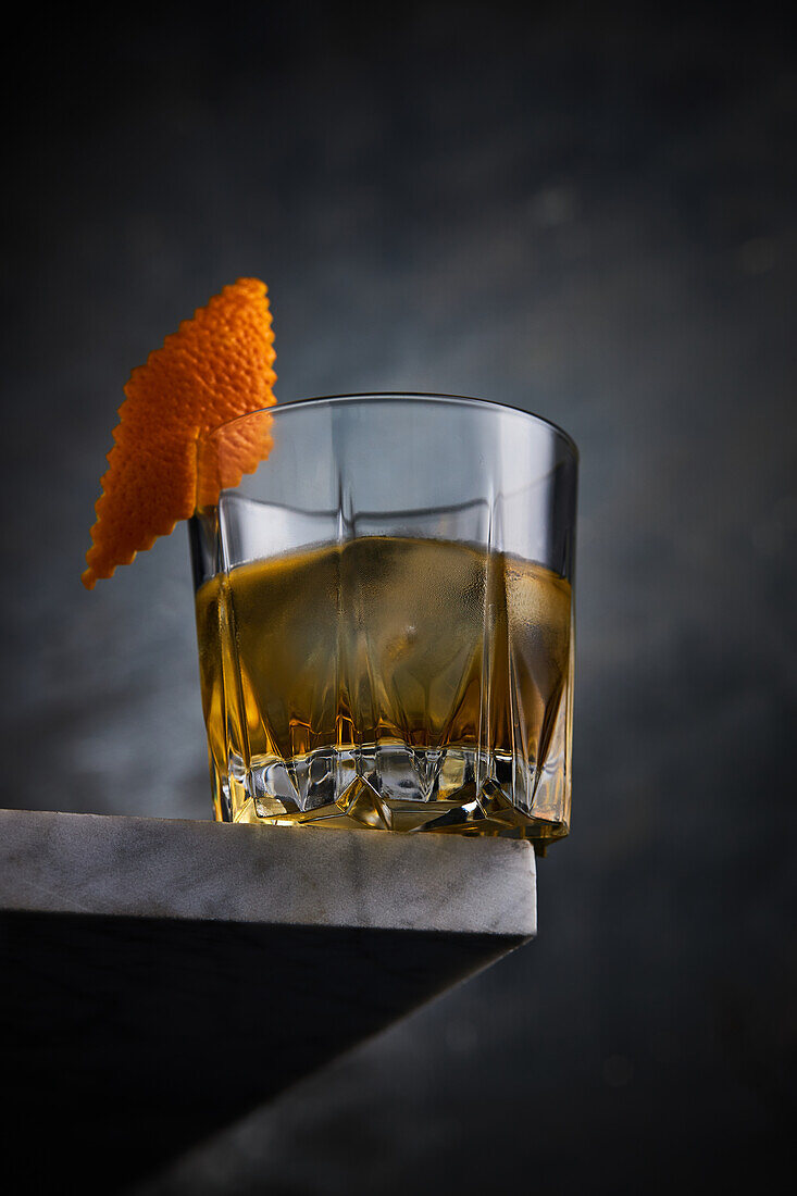 Glass full of alcoholic Scotch whiskey decorated with orange peel placed on corner of table against dark background in room