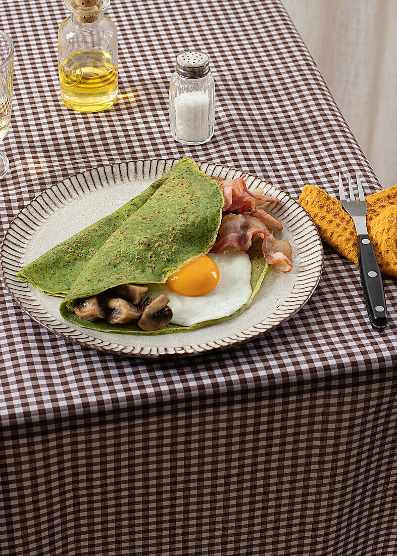 Homemade breakfast of spinach pancakes with bacon, egg and mushrooms served on a white plate with a salt and oil shaker on a checkered tablecloth.