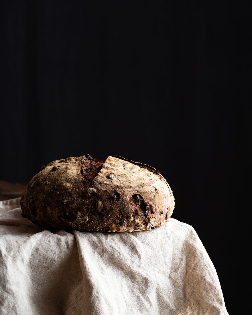 Freshly baked sourdough bread loaf with walnuts and raising placed on white linen on black background
