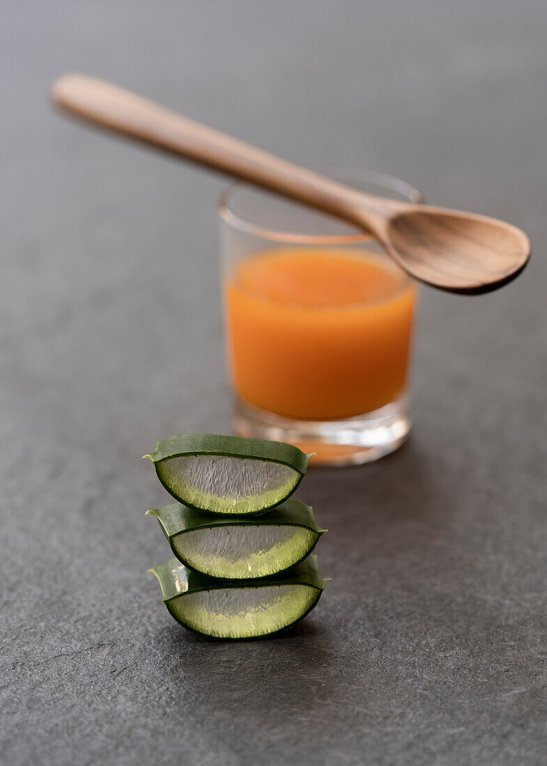 Cut pieces of aloe vera placed on gray table with glass of orange juice and wooden spoon