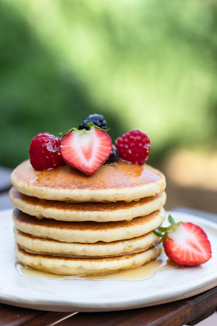 Stapel leckerer Pfannkuchen mit frisch geschnittenen Erdbeeren und Himbeeren auf weißem Teller mit Honig vor unscharfem Hintergrund
