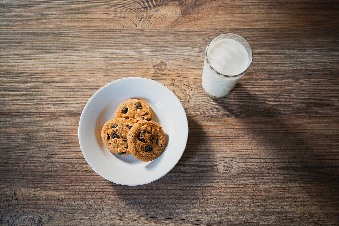 Blick von oben auf einen Teller mit leckeren Haferflockenkeksen mit Schokoladenstückchen vor einem Glas Milch auf einem Holztisch