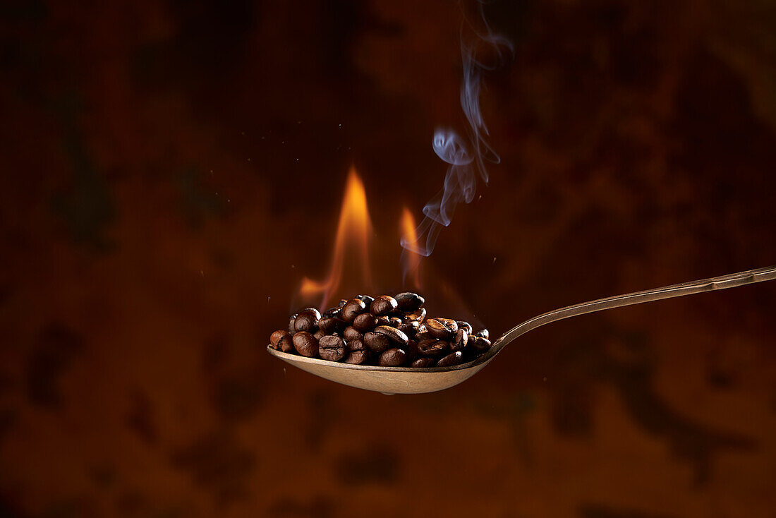 Closeup of spoon full of heap of roasted coffee beans with fire on brown background