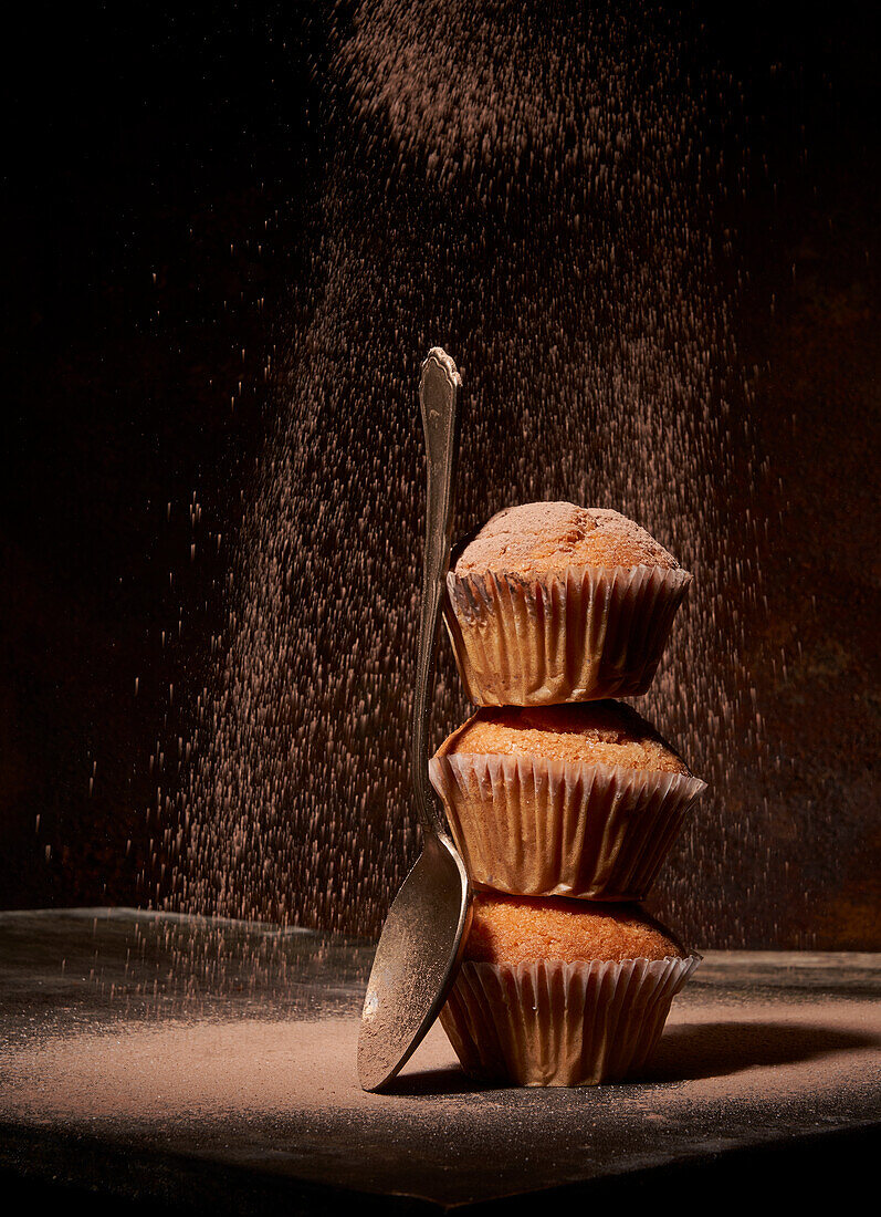 White powdered sugar sprinkling on tasty sweet baked muffins stacked on top of each other near spoon on black background