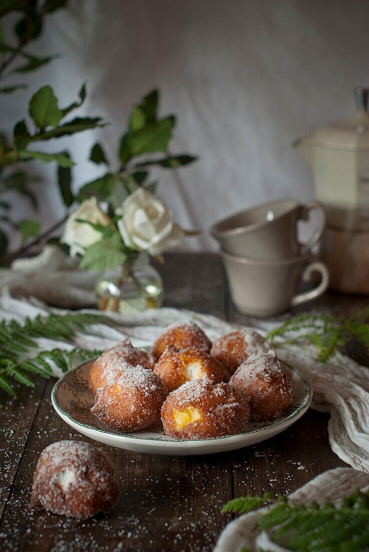 Hausgemachte Puddingcremekrapfen mit Zucker bestreut auf rustikalem Holztisch mit Tischtuch und Blätterdekoration