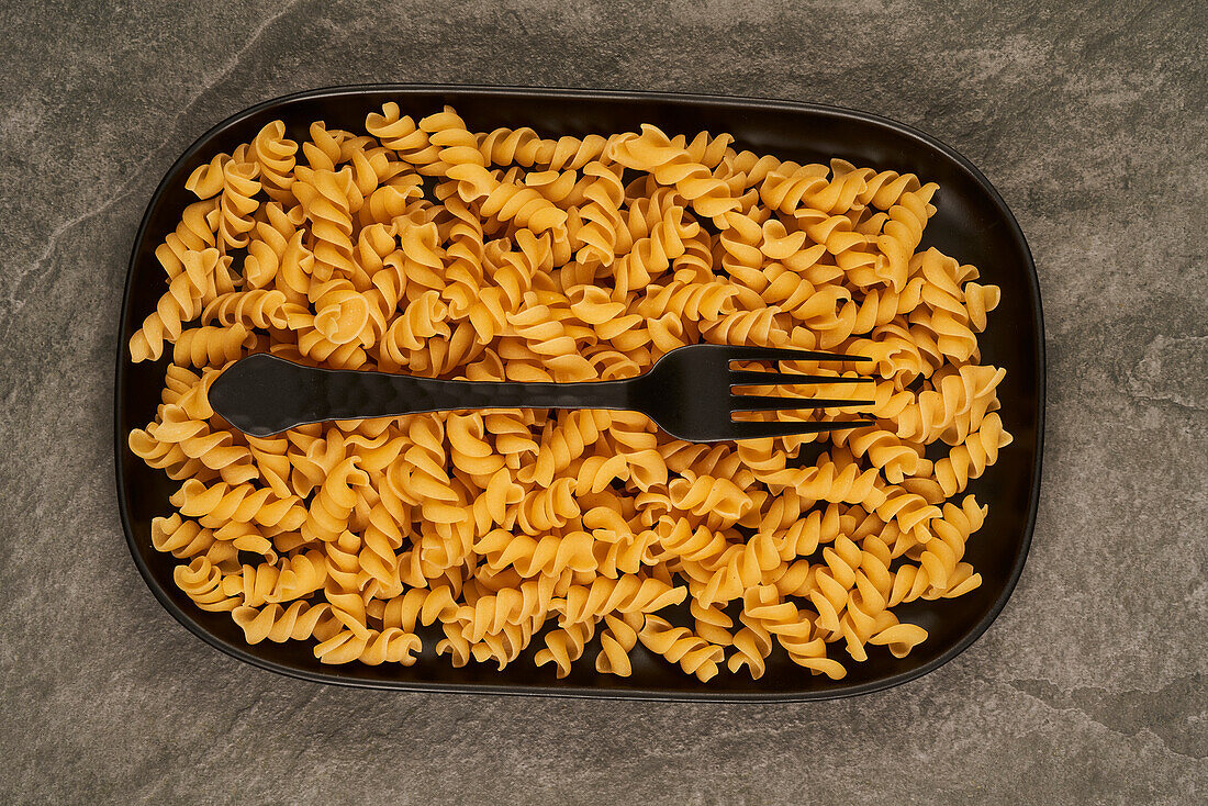 Schwarze Gabel von oben neben ungekochten Fusilli-Nudeln auf einem Tablett auf dem Tisch