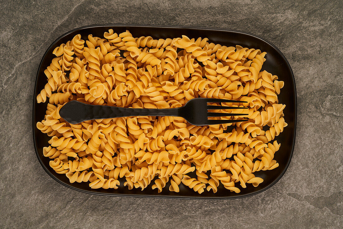 Top view of black fork placed near uncooked fusilli pasta on tray on table