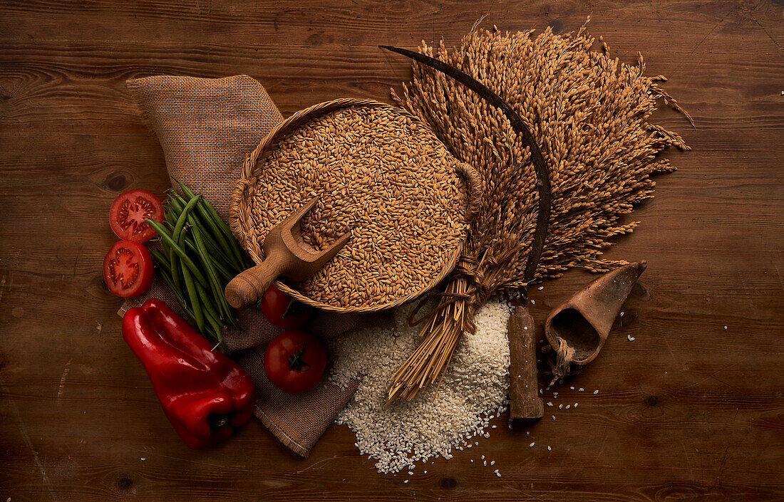 From above rustic composition of harvesting sickle on bunch of wheat ears near bowl full of golden corns and fresh vegetables on burlap on wooden table