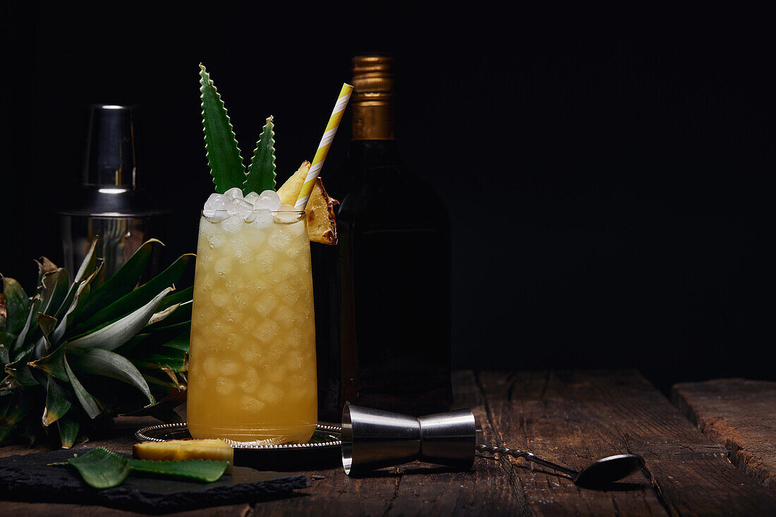 Glas eines alkoholischen Cocktails mit Ananasstück und Blättern mit Papierstrohhalm auf einem Tablett neben einem Shaker und einer Flasche mit Schnapsglas auf einem Tisch mit schwarzem Hintergrund