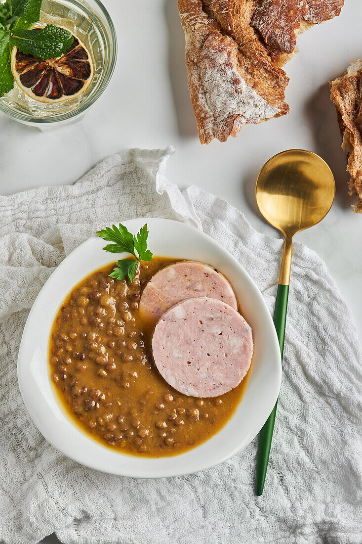 Draufsicht auf einen Löffel neben einer Schüssel mit leckerer Linsensuppe, serviert mit Wurstscheiben und einem Blatt Petersilie auf einem Marmortisch mit grauer Serviette