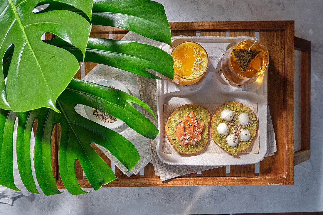 From above of delicious avocado toasts with salmon and burrata cheese served on table with glasses of fresh juice and herbal tea placed near exotic Monstera deliciosa plant