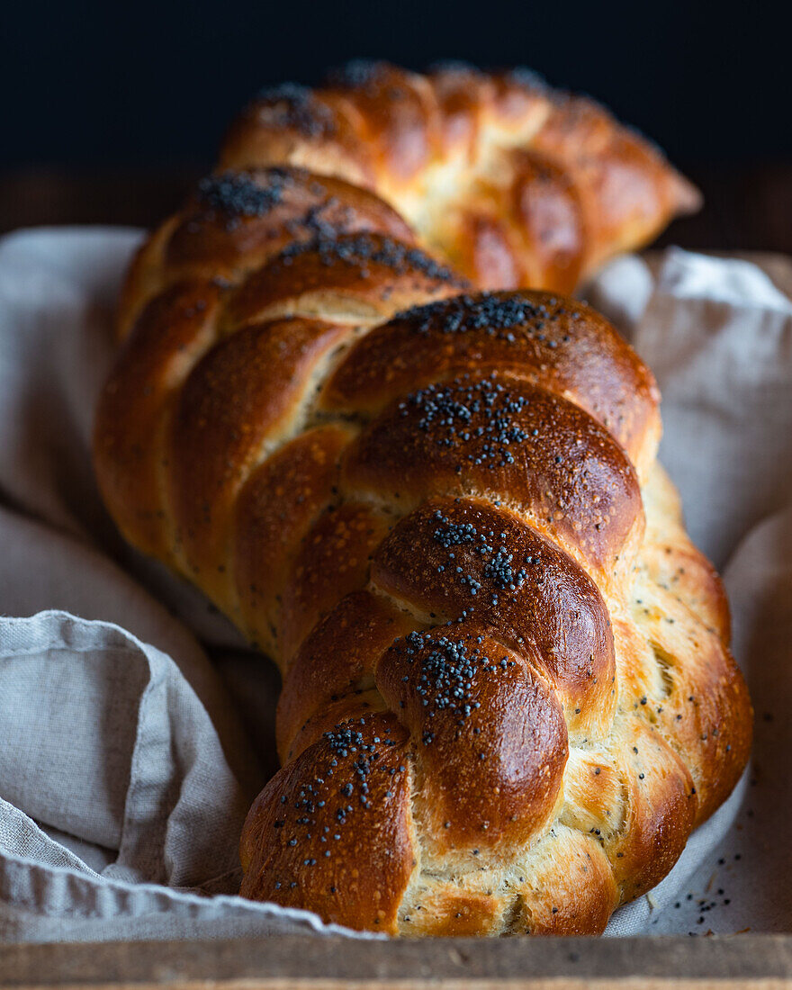 Served golden brown bread braids challah with poppy seeds on white napkin in wooden box