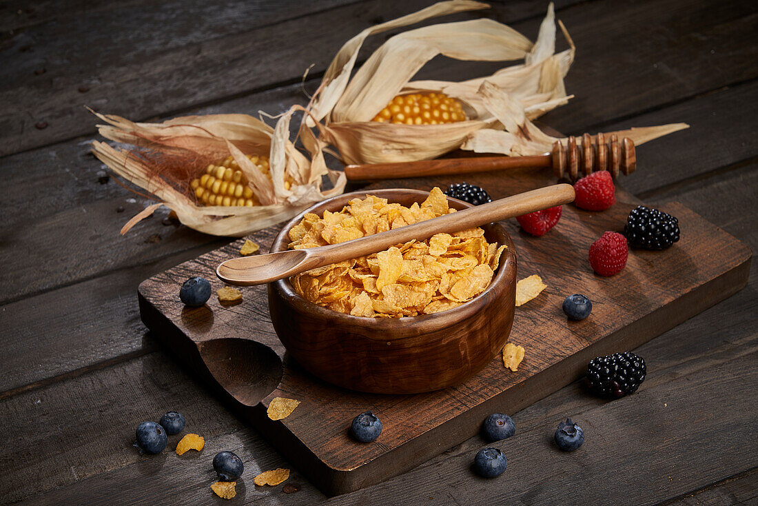 Schüssel mit leckeren süßen Cornflakes mit Löffel auf hölzernem Schneidebrett auf Tisch mit Maiskolben und Beeren in heller Küche