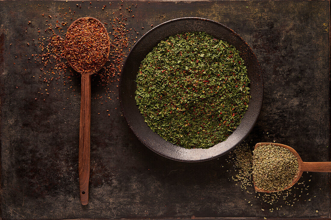 Top view of bowl with green dried herbs and spoon with ground sun dried tomato powder with artificial wooden hand on black background