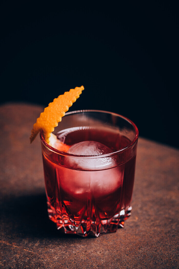 Glass of refreshing Negroni cocktail with bitter flavor and ice garnished with orange peel and served on couch arm in dark room