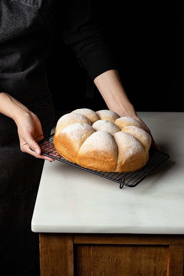 High angle of crop unrecognizable baker standing at counter with fresh bread buns placed on baking pan in kitchen
