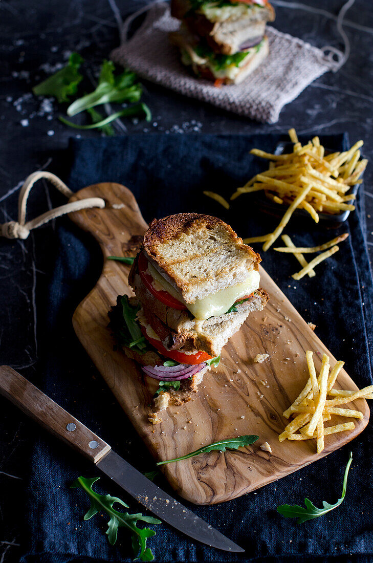 Draufsicht auf ein leckeres Sandwich mit Tomate, Zwiebel, Käse und Fleisch auf einem Holzbrett mit Pommes frites und Messer auf einem blauen Tisch