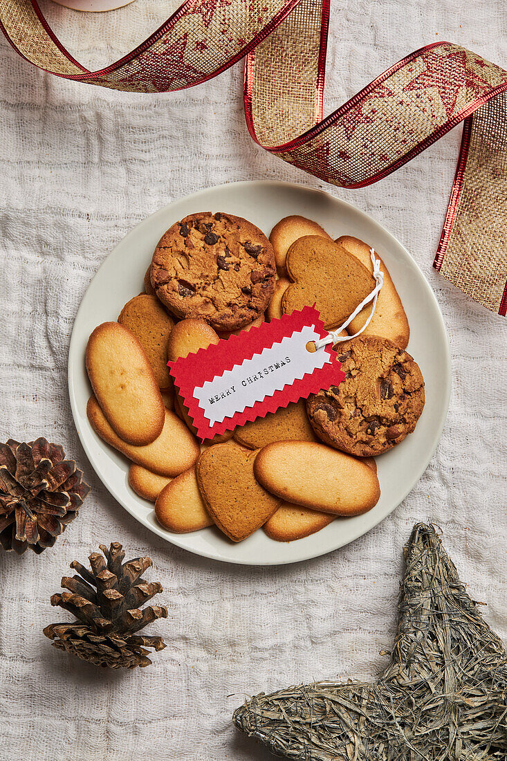 Teller mit verschiedenen süßen Keksen und Geschenkanhänger auf einem Tisch mit Weihnachtsdekoration von oben