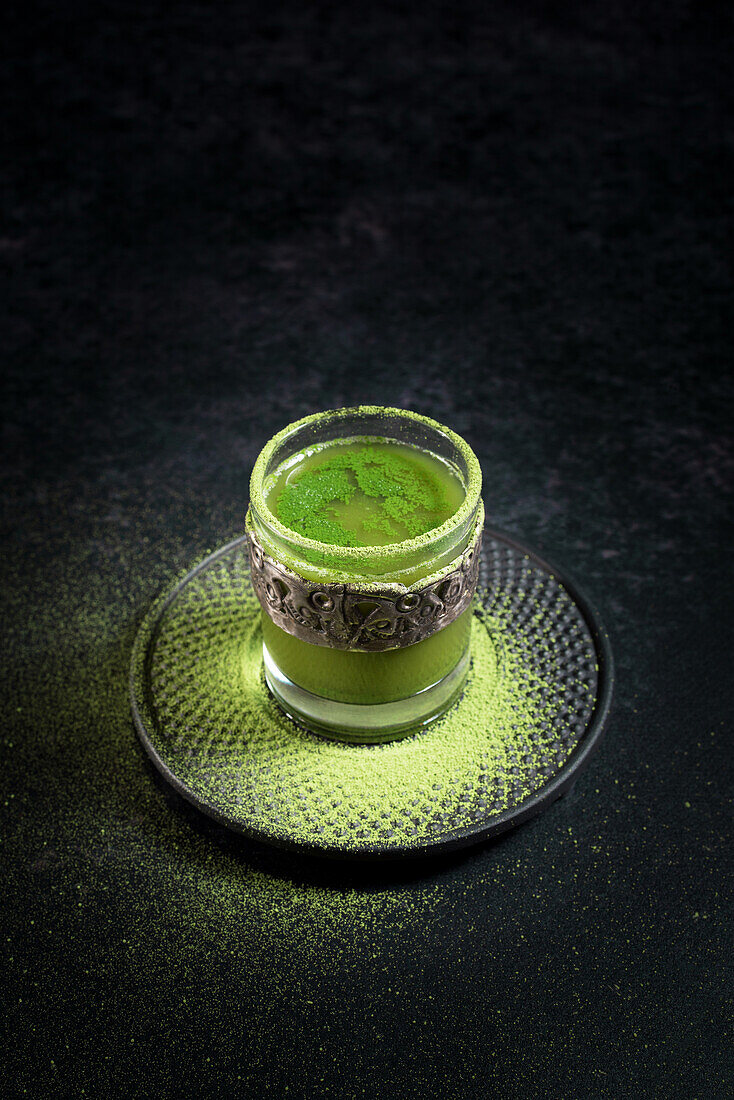 From above of healthy herbal green matcha tea served in glass cup with metal decoration on saucer sprinkled with powder on black table