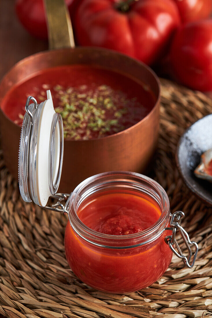 Top view of pan and container with delicious homemade tomato sauce sprinkled with oregano placed on woven mat on wooden table in kitchen