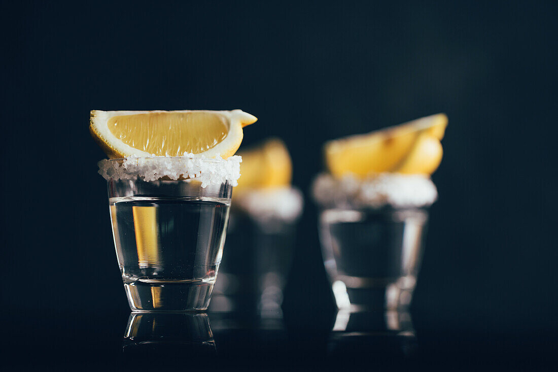 Tequila shots with salt and lemon placed on reflective surface against dark background