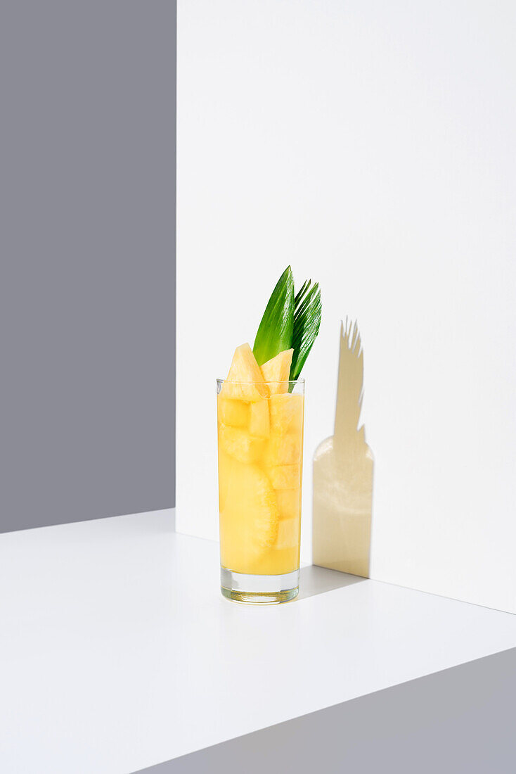 Glass full of cold pineapple juice with green leaves and straw placed on sunlit table against white background