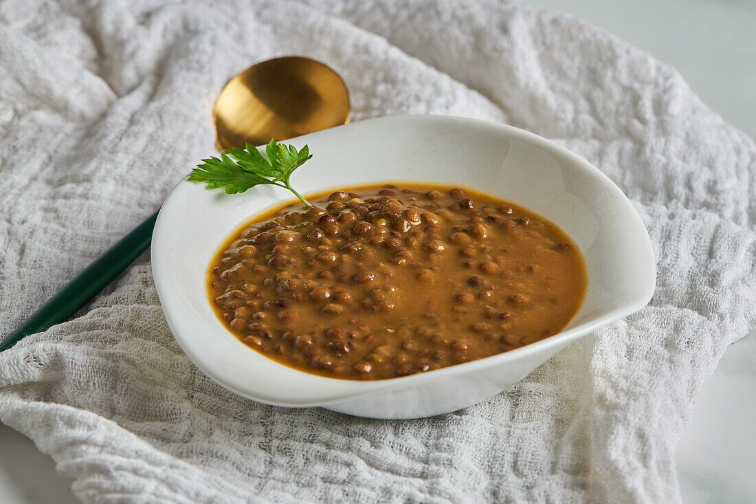 From above of white bowl of delicious lentil soup garnished with parsley placed on napkin near spoon