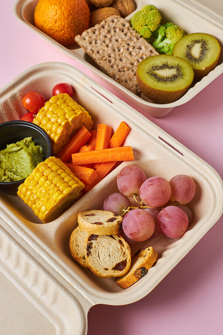 From above of lunch boxes with healthy food including crackers carrot sticks grapes cherry tomatoes with kiwi broccoli walnut and tangerine on pink background