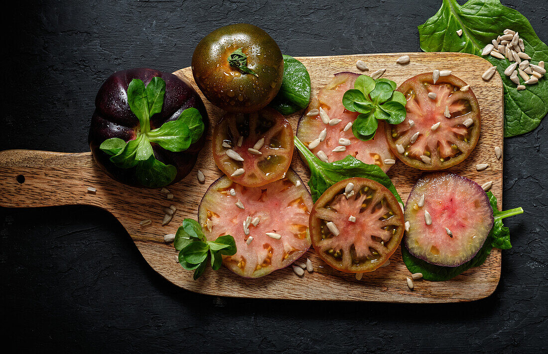 Draufsicht auf frische, reife, in Scheiben geschnittene schwarze Tomaten und grüne Minzstängel auf einem hölzernen Schneidebrett auf schwarzem Hintergrund