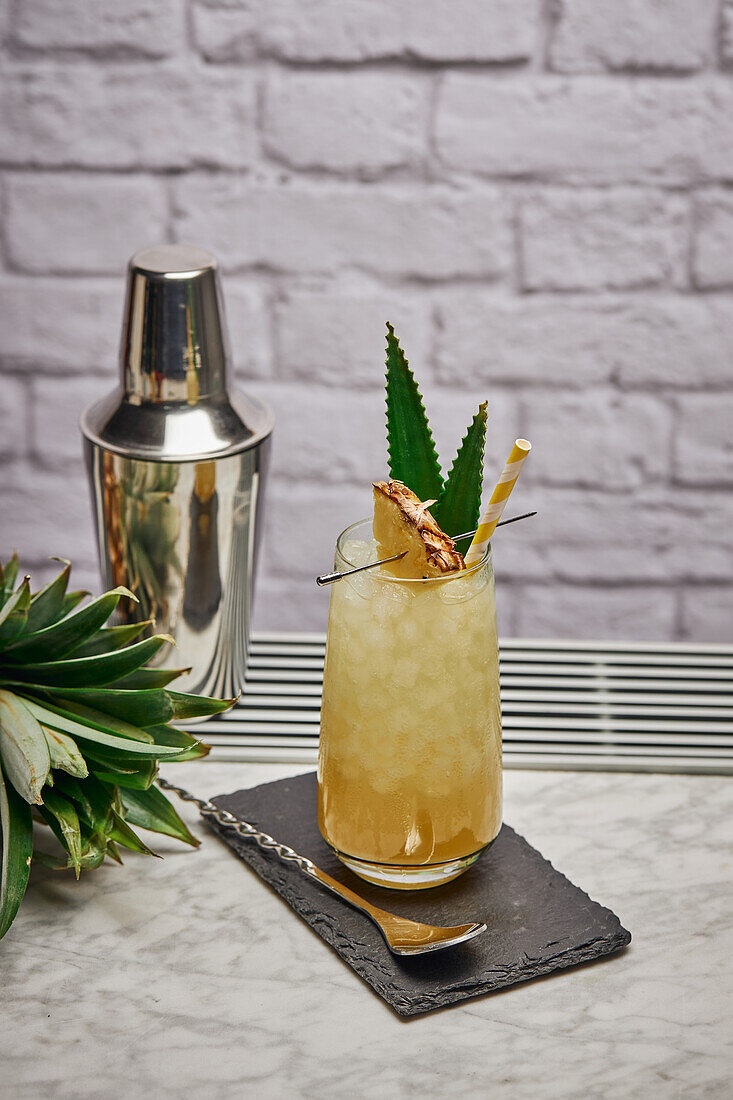 Yellow cocktail in glass garnished with pineapple piece and green leaves with paper straw placed on slate coaster with bar spoon
