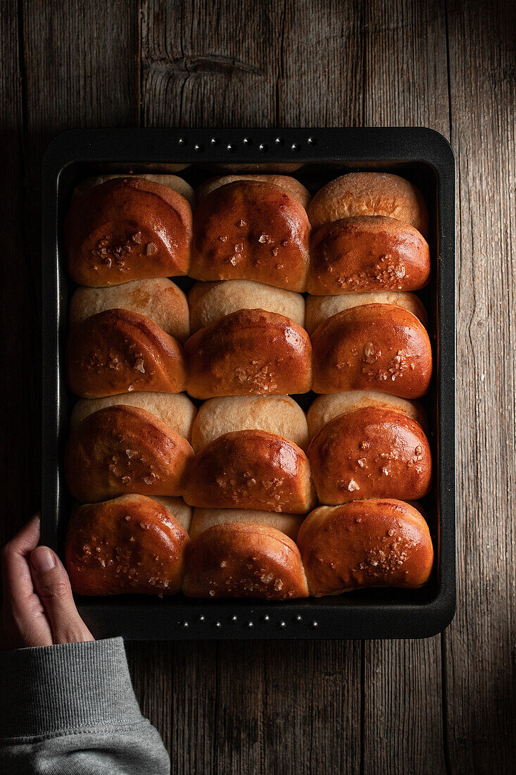 Blick von oben auf einen gesichtslosen Erntekoch, der eine Backform mit appetitlichen weichen Brötchen auf einem Holztisch hält