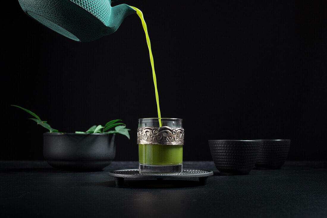 Gesunder japanischer Matcha-Tee, der während einer Teezeremonie aus einer grünen Teekanne in eine Glastasse mit Metallornamenten gegossen wird, vor schwarzem Hintergrund