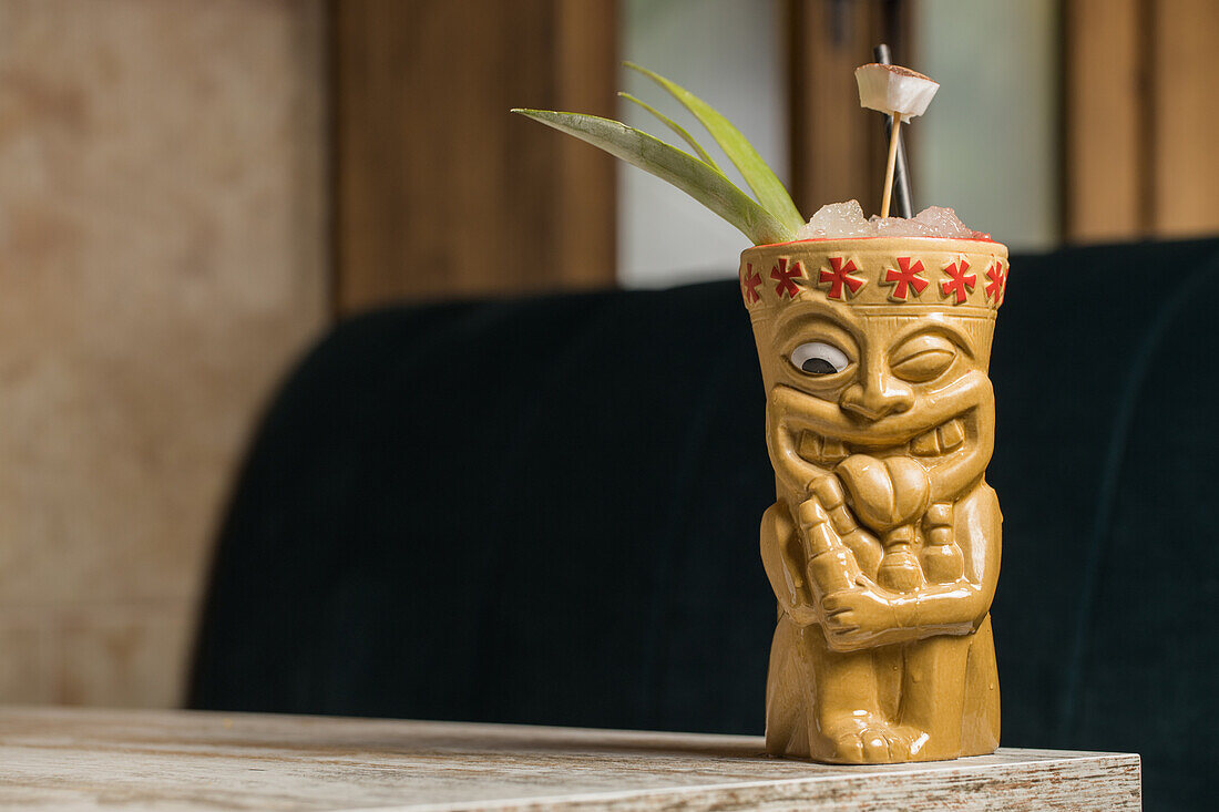 Polynesischer Tiki-Becher mit kaltem alkoholischem Getränk, dekoriert mit Strohhalm und grünen Ananasblättern, auf einem Holztisch