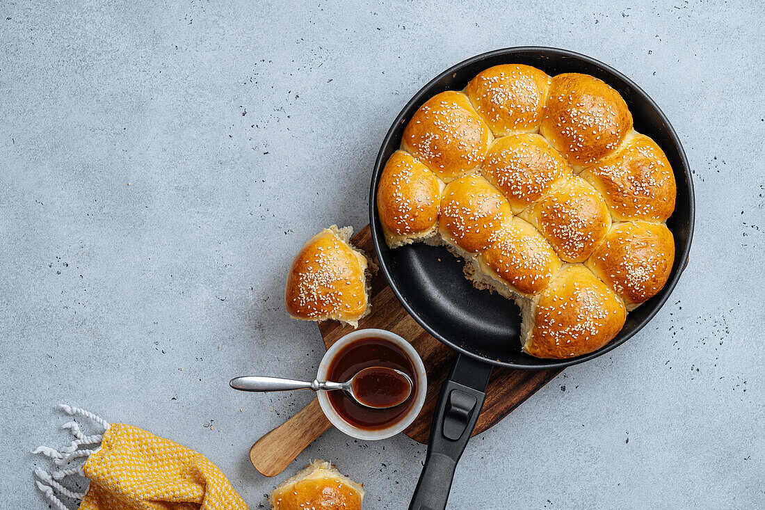 From above of tasty pull apart bread rolls with sesame seeds in frying pan served on wooden cutting board with sweet sauce