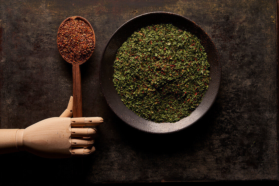 Top view of bowl with green dried herbs and spoon with ground sun dried tomato powder with artificial wooden hand on black background